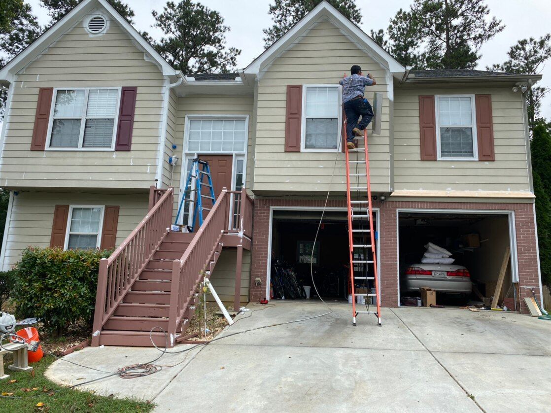 Two workers on ladders painting the front of a beige house with a staircase and open garage.