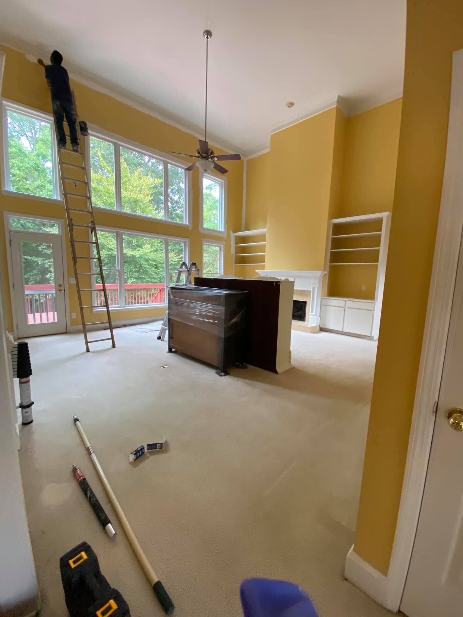 High-ceiling living room under renovation with ladder, tools, and large windows.