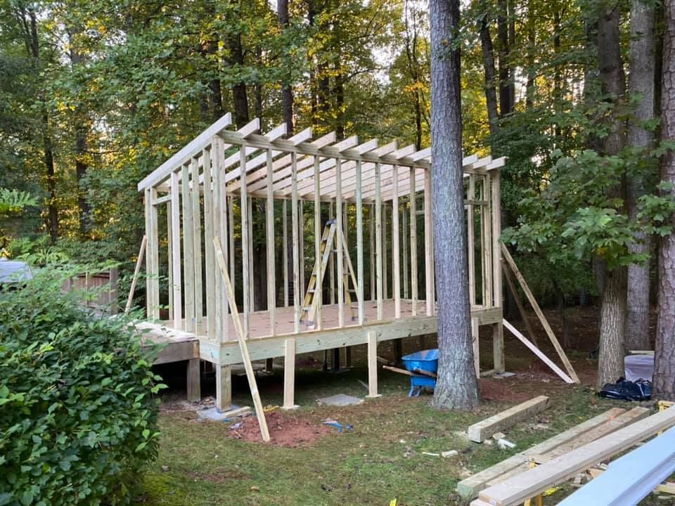 Partially framed wooden shed on a raised platform in a wooded yard, with lumber stacked nearby.