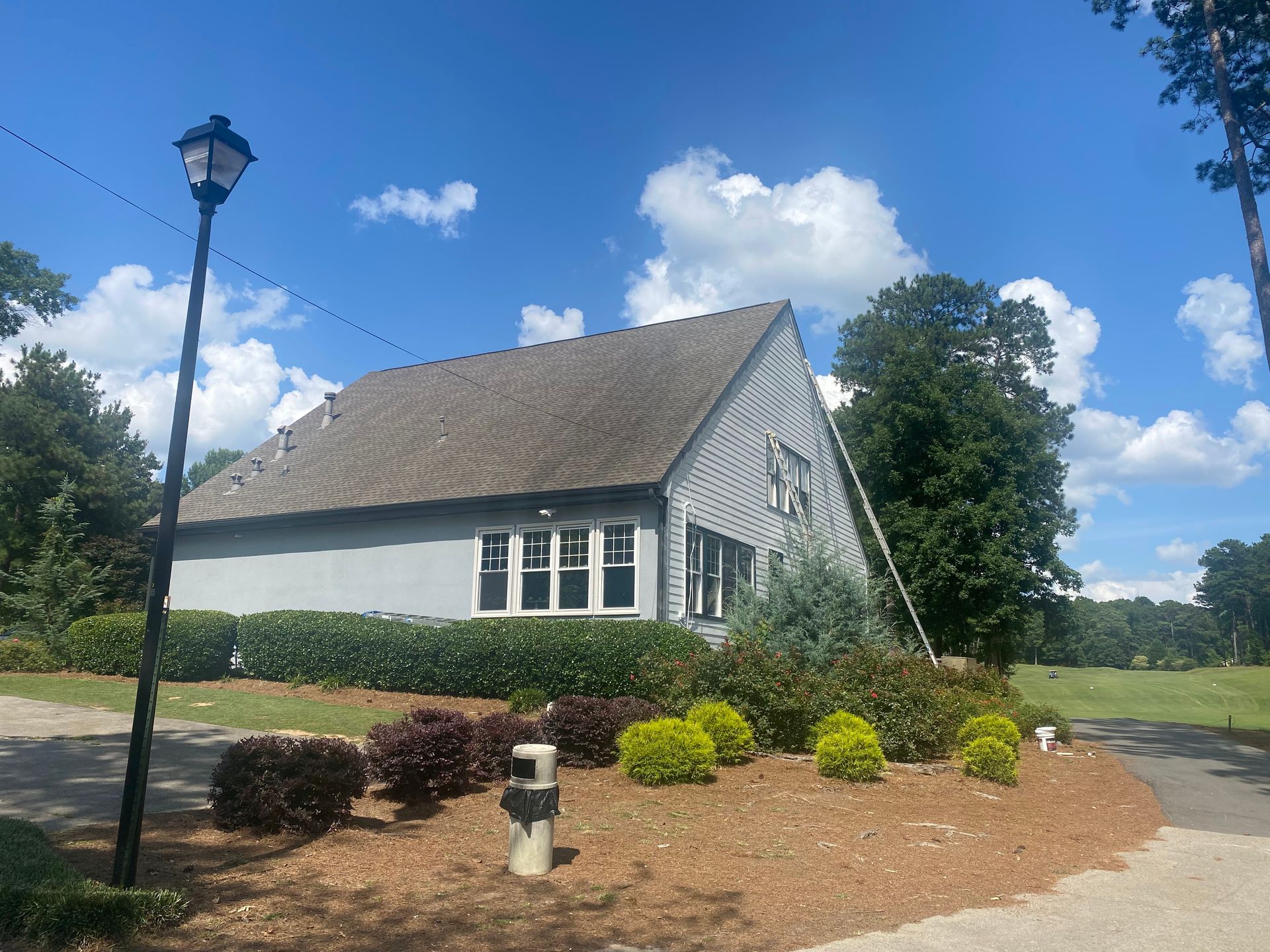 White building with gray roof, lamppost, and landscaped shrubs under a blue sky.