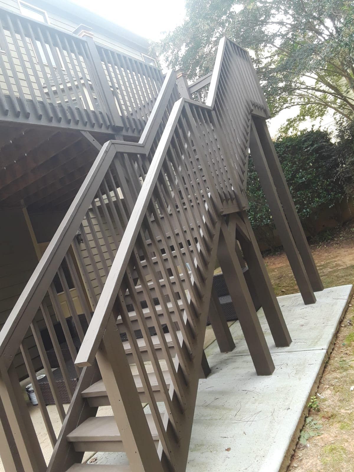 Wooden outdoor staircase with railings leading up a deck beside a concrete path and trees