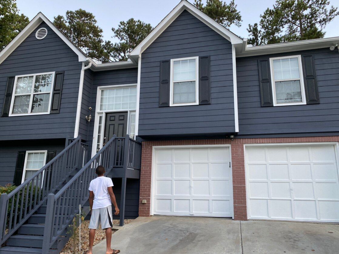 Blue two-story house with white trim and garage; person stands on front steps.