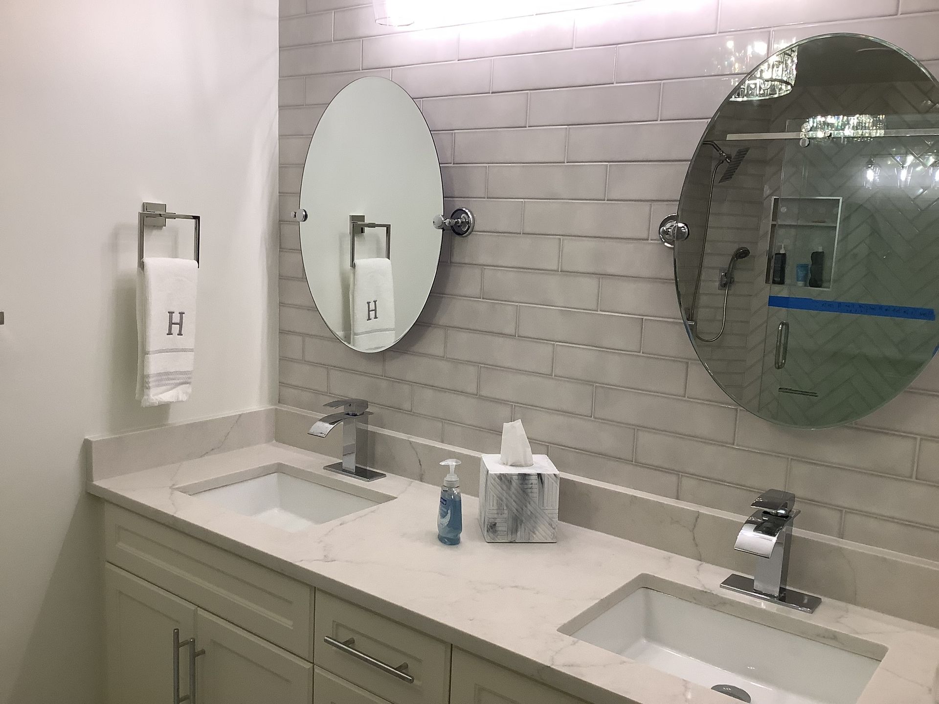 Modern restroom with dual sinks, oval mirrors, tiled wall, and soap dispensers