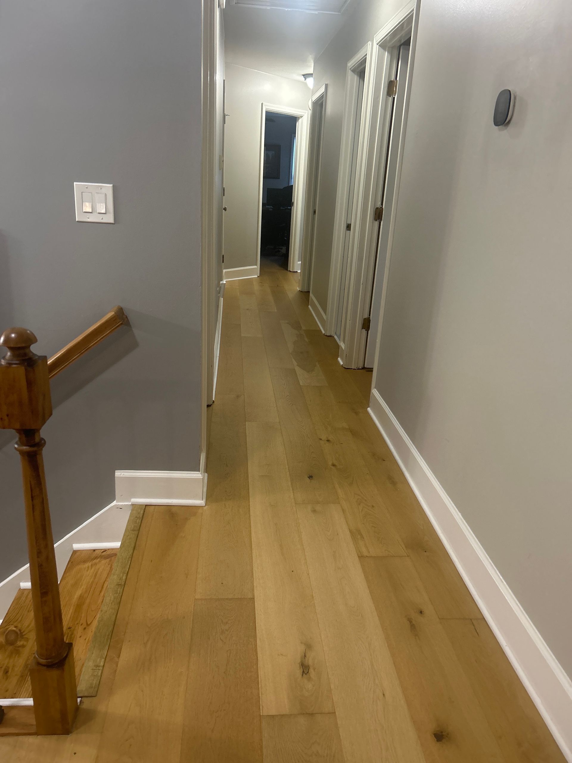 Narrow hallway with light wood floor, gray walls, staircase railing, and closet doors on the right.