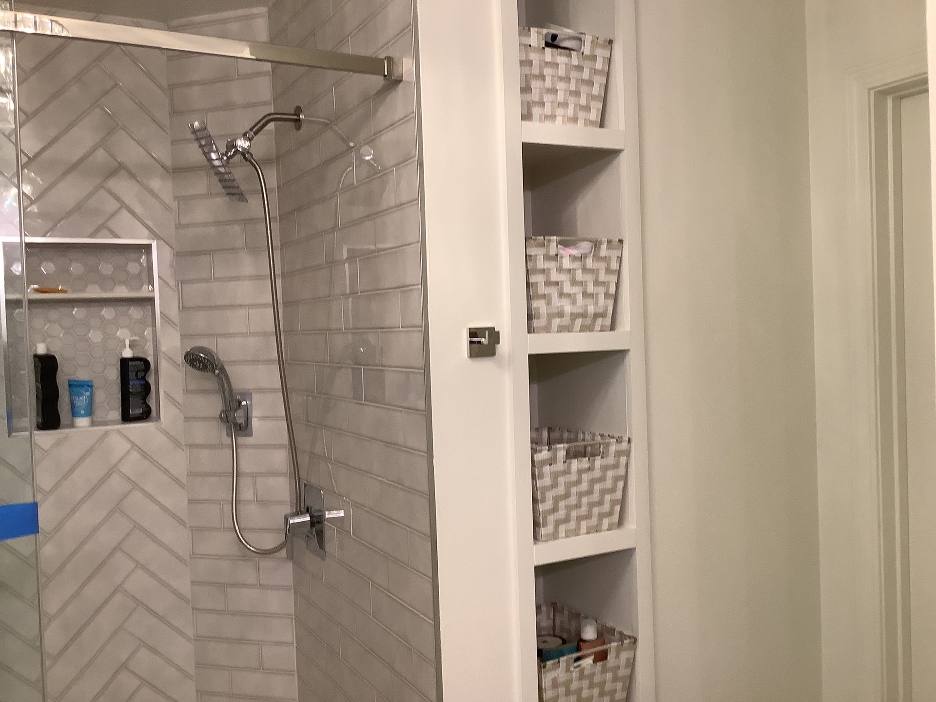 White tiled bathroom shower with chrome fixtures, built-in shelf, and woven storage baskets.