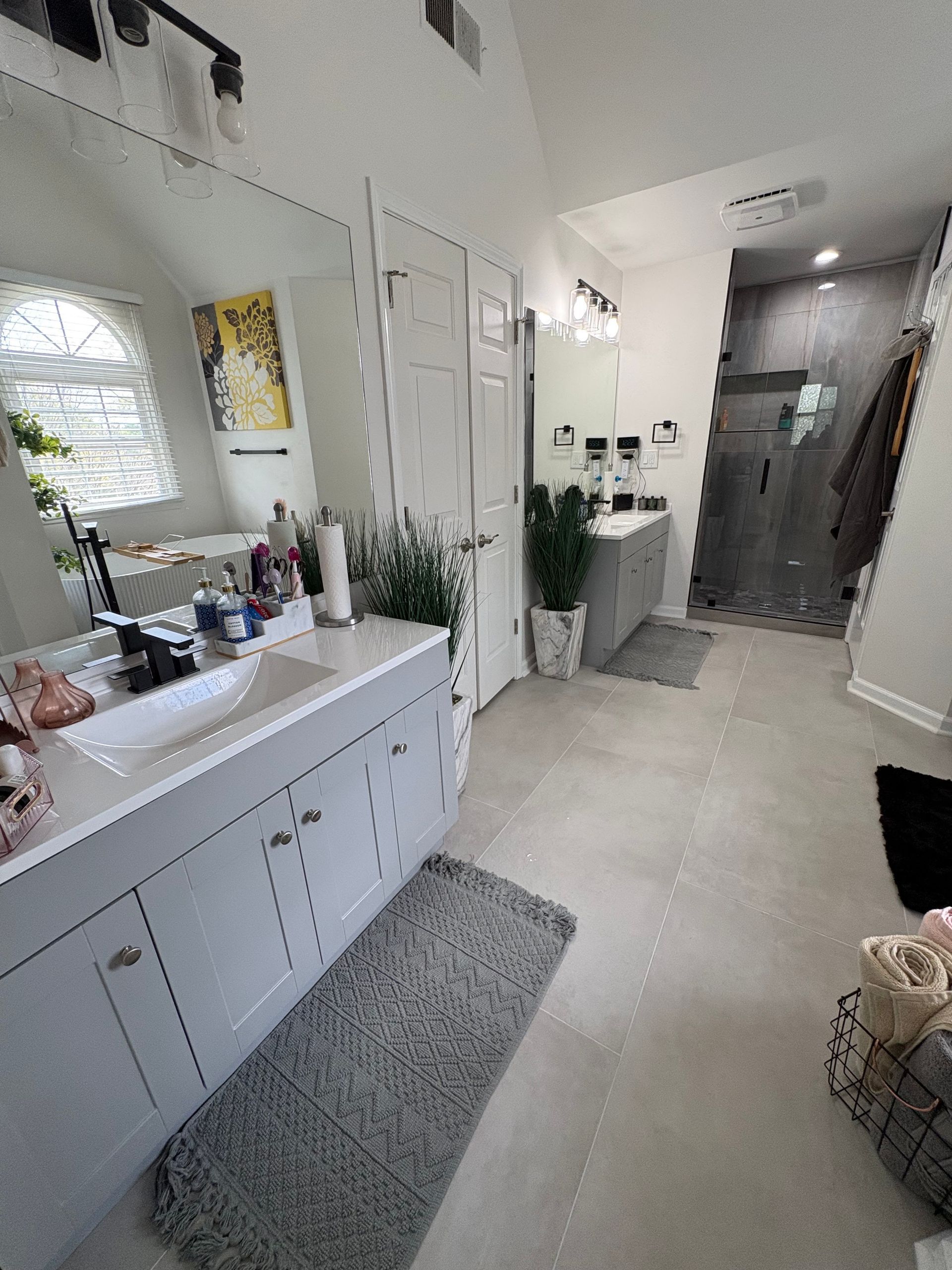 Bright white bathroom with a double sink, large mirror, rug, plants, and a glass shower at the end of the hall
