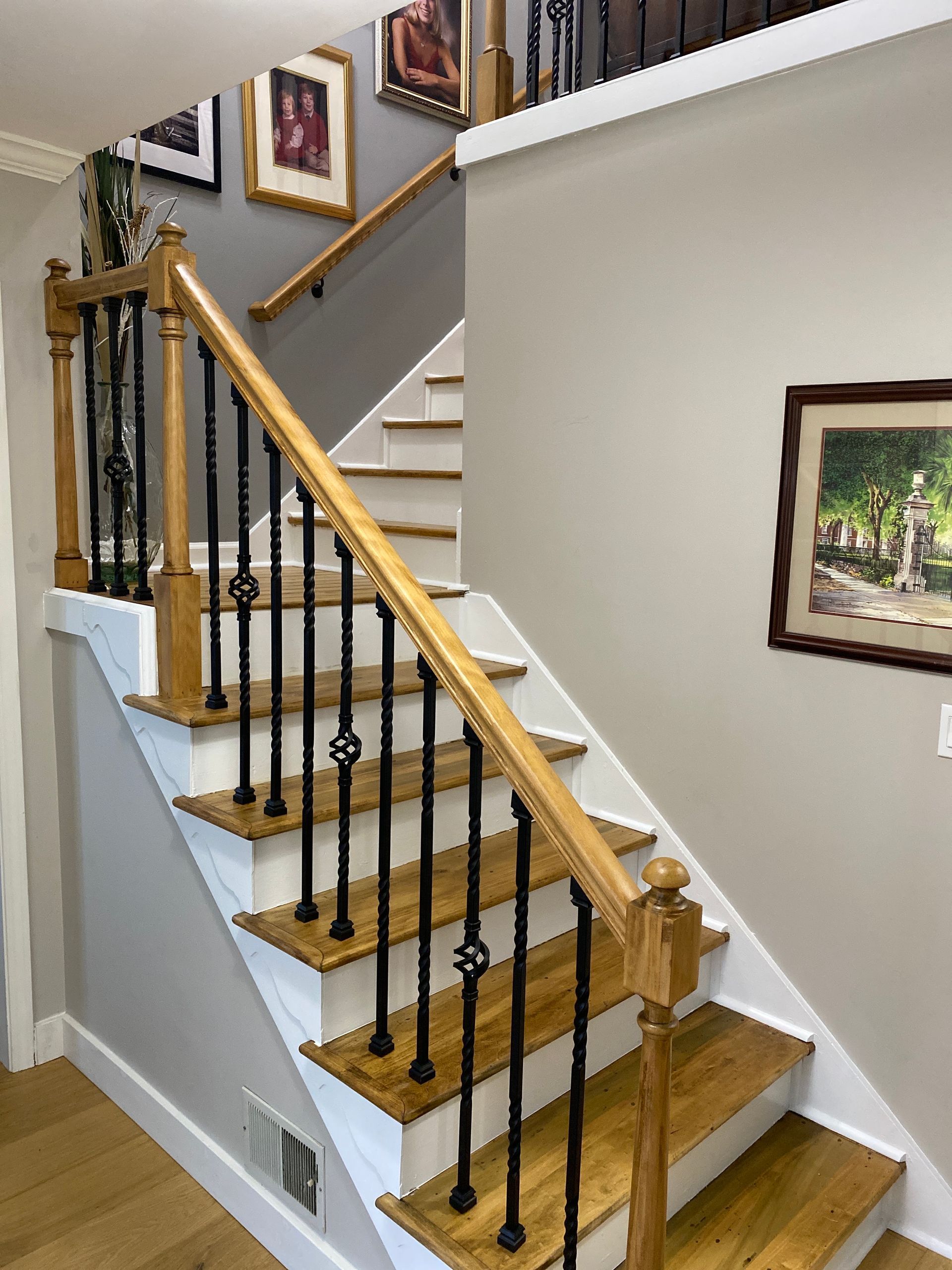 Interior staircase with wooden steps, black iron balusters, and framed wall art along a white hallway.