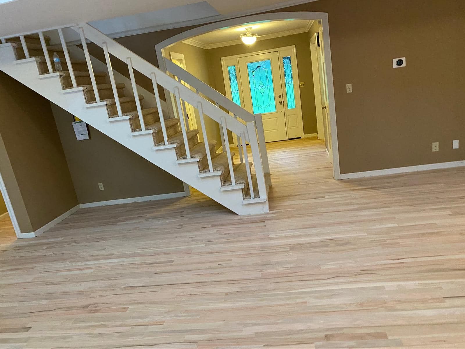 Empty living room with light wood floors, tan walls, and a white staircase leading to a bright hallway.