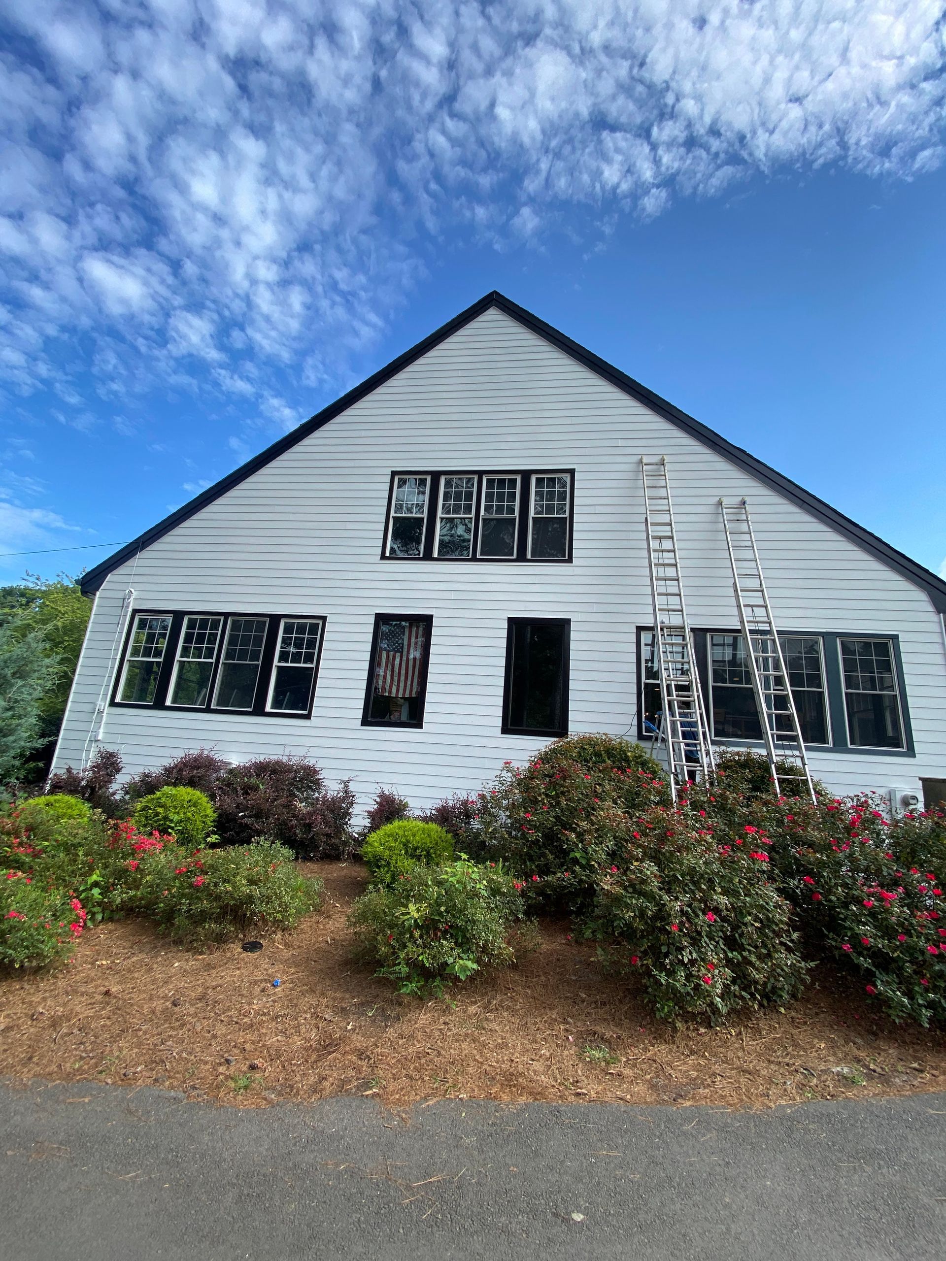 White clapboard house with black-trimmed windows, garden shrubs, and a ladder under a blue sky