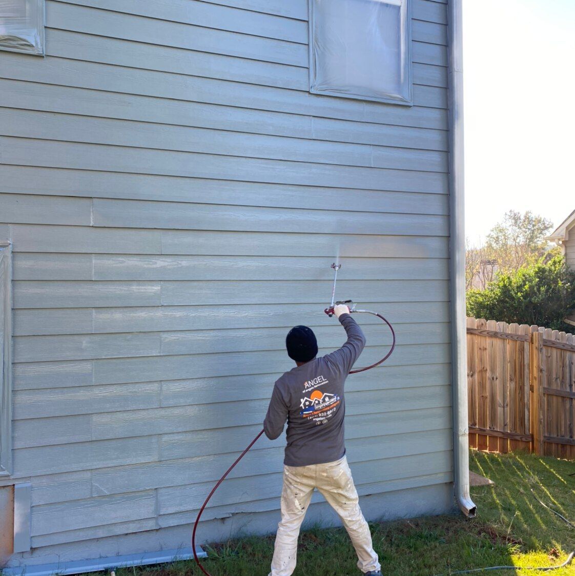 Person power-washing a blue-gray house exterior beside a fenced yard.