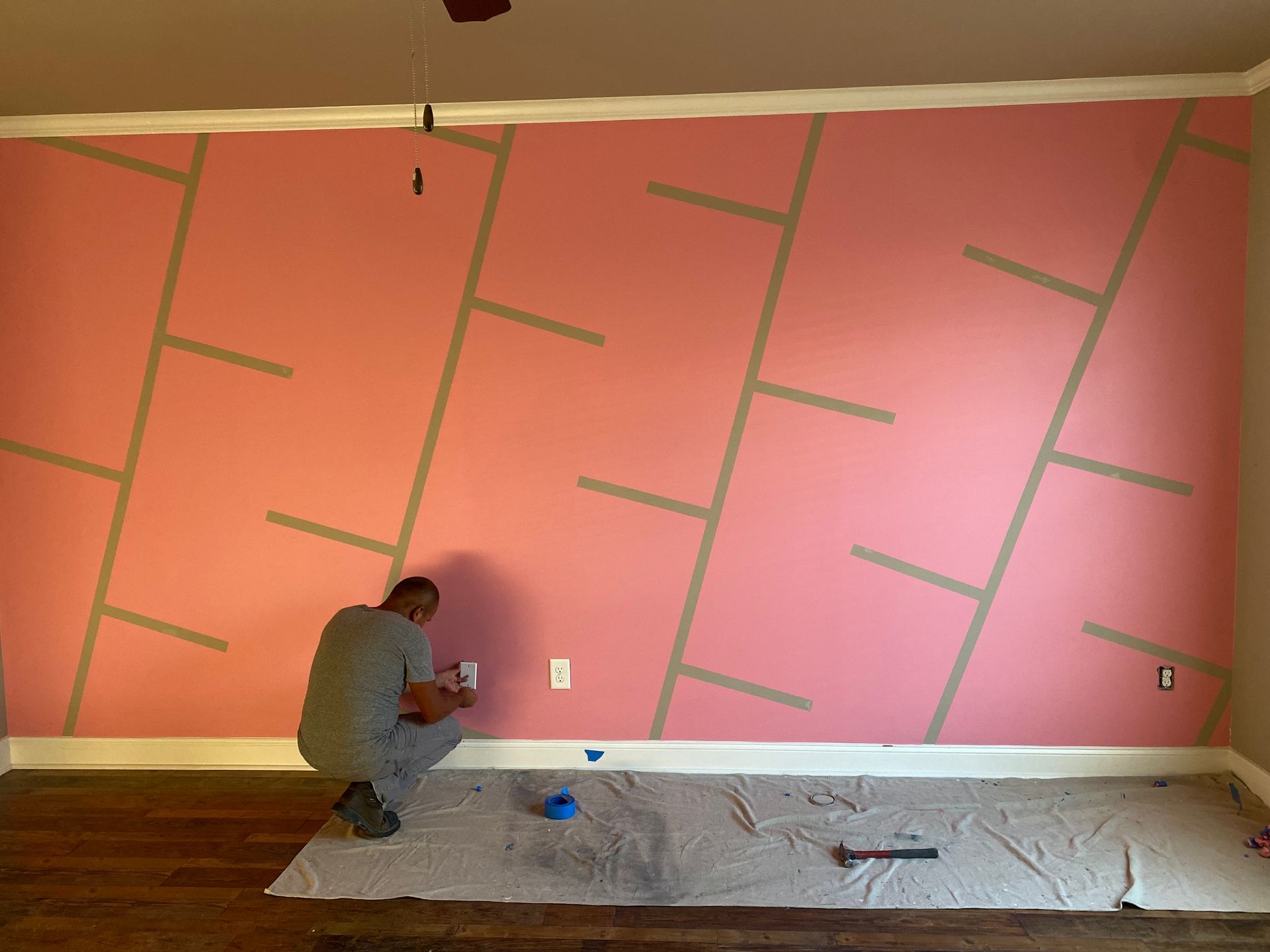 Person kneeling and taping off a pink wall for painting in a room with plastic-covered floor