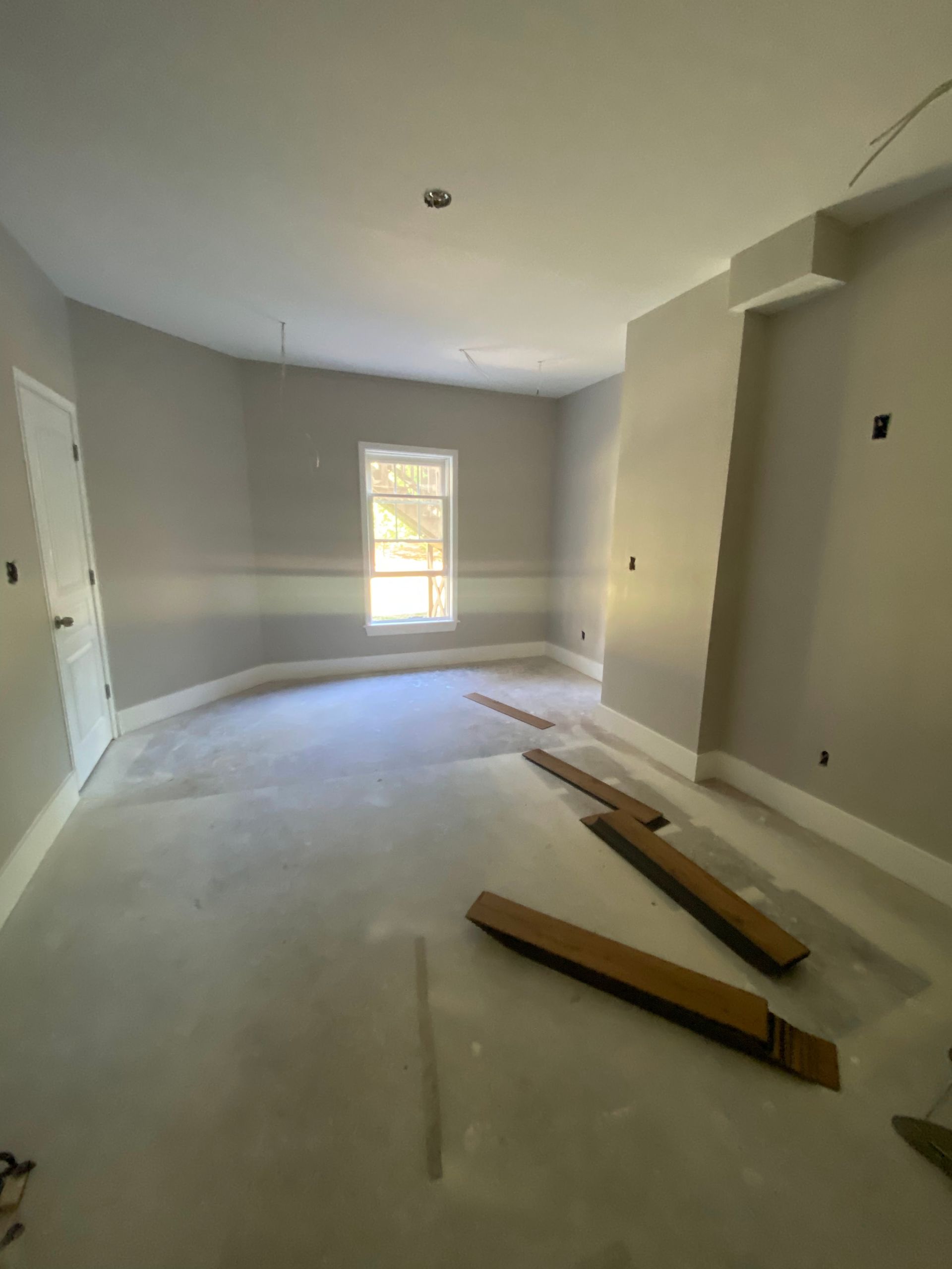 Empty unfinished room with beige walls, white ceiling, carpeted floor, and construction materials near a window.
