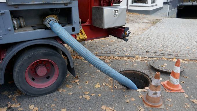 Blue hose from truck to ground near orange traffic cones on a leaf-strewn street.