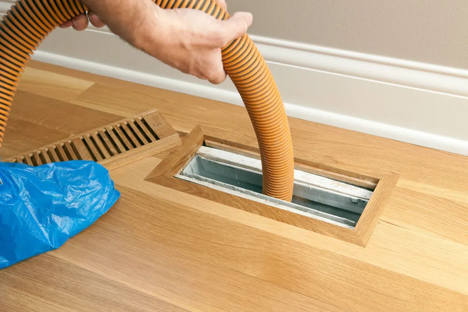 Hand vacuum hose cleaning floor vent in a hardwood floor, with a blue bag nearby