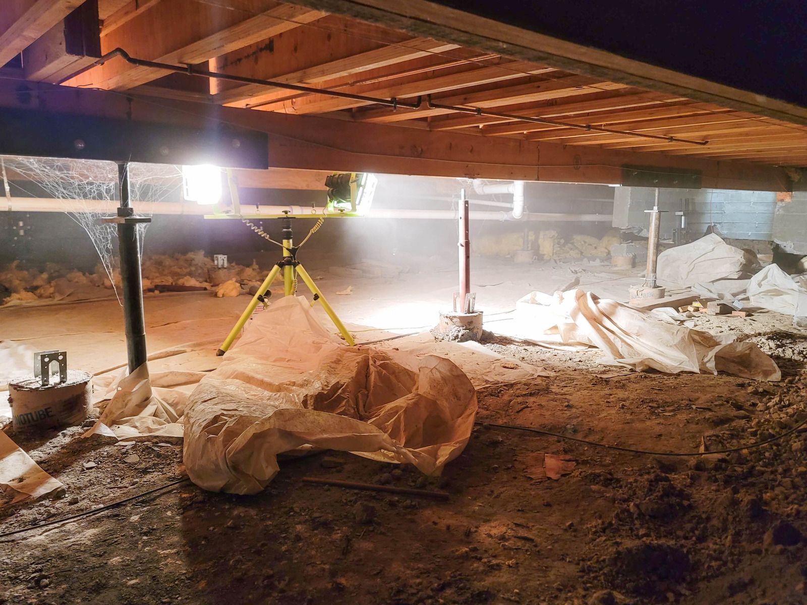 Crawlspace under a house with yellow support jacks, exposed beams, and debris-covered ground.
