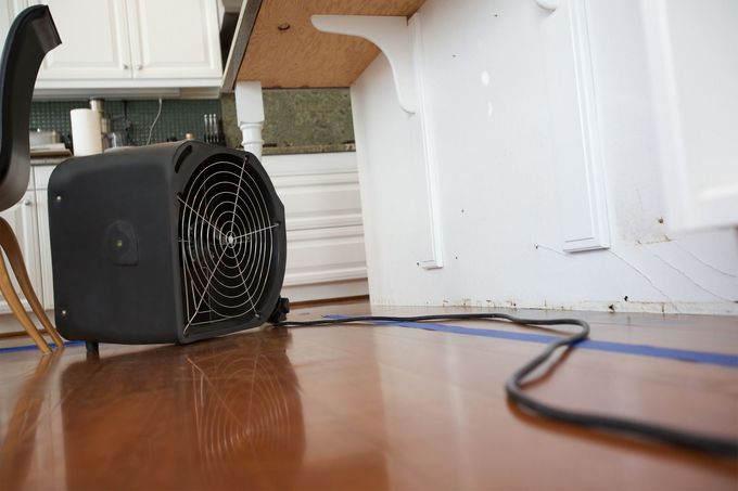 Black fan on a hardwood floor beside white cabinets and a loose cord in a kitchen.