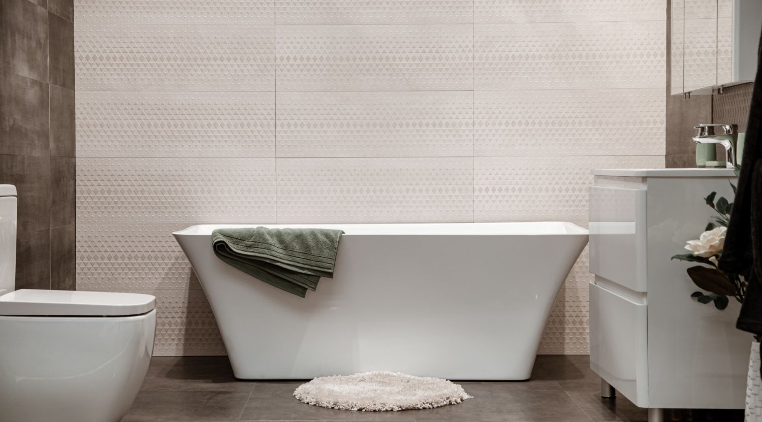 Modern bathroom with white bathtub, toilet, vanity, and beige tiled wall; green towel draped over tub.