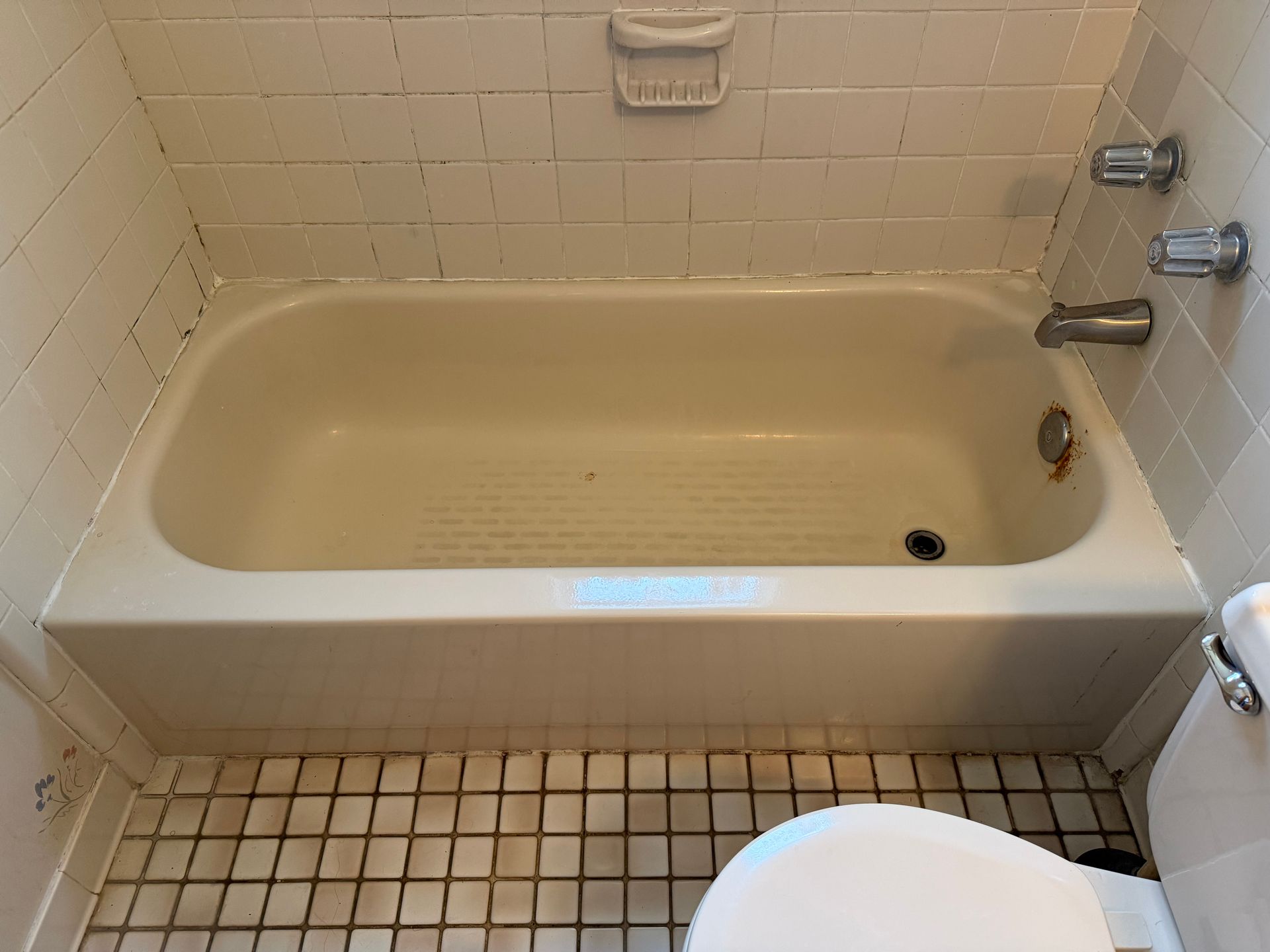 Empty beige bathtub in a small tiled bathroom with a toilet visible in the foreground