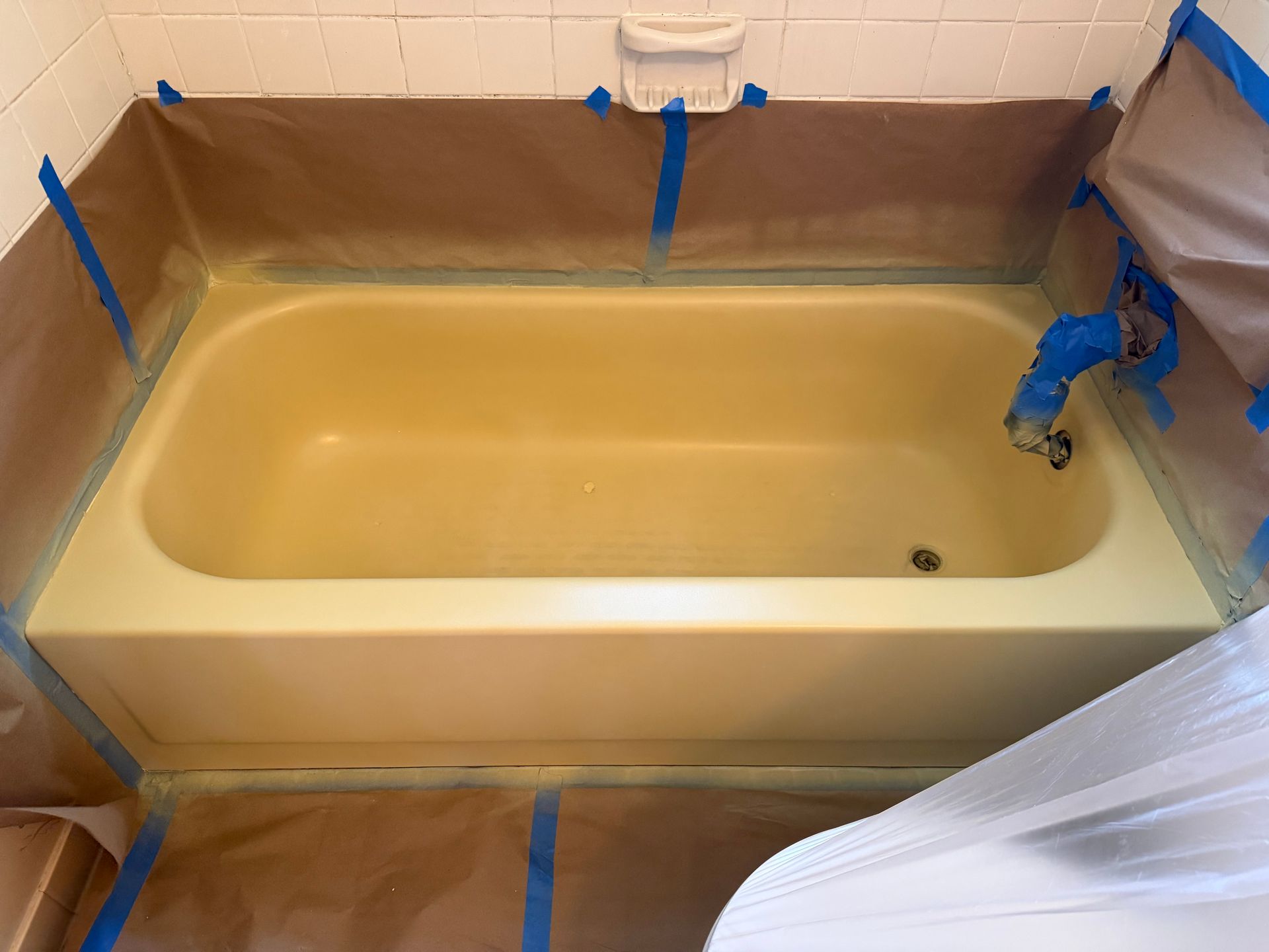 Bathtub with tan liner, blue tape, and plastic sheeting in a bathroom under renovation