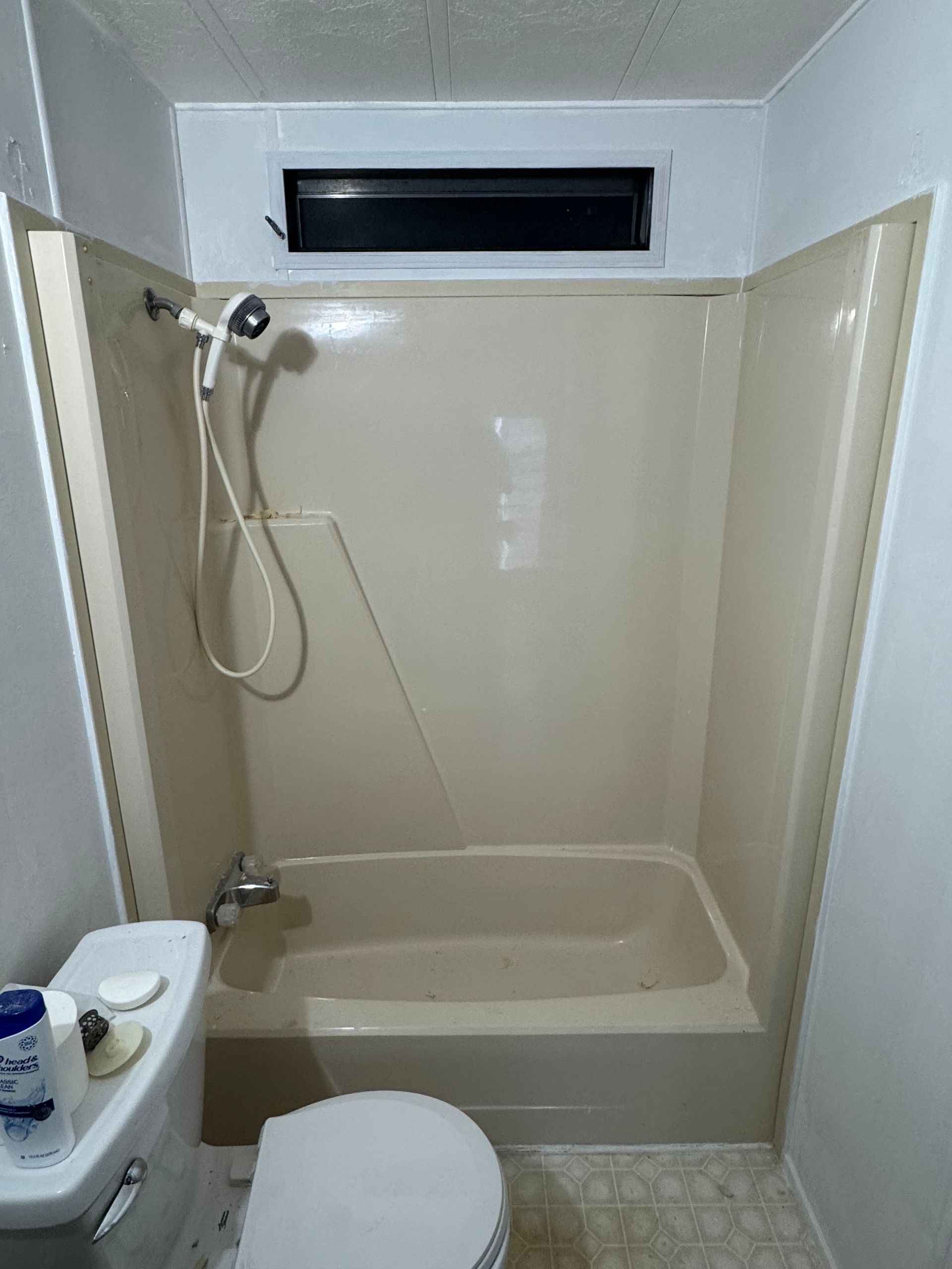 Small bathroom with beige tub, showerhead, toilet, and a narrow frosted window above the tub.