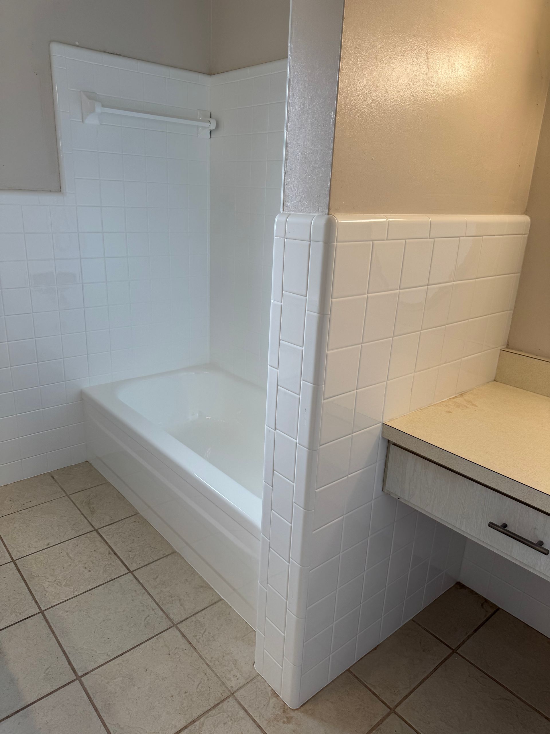 Small bathroom with white tub, tiled shower wall, and beige sink counter divided by a half wall.