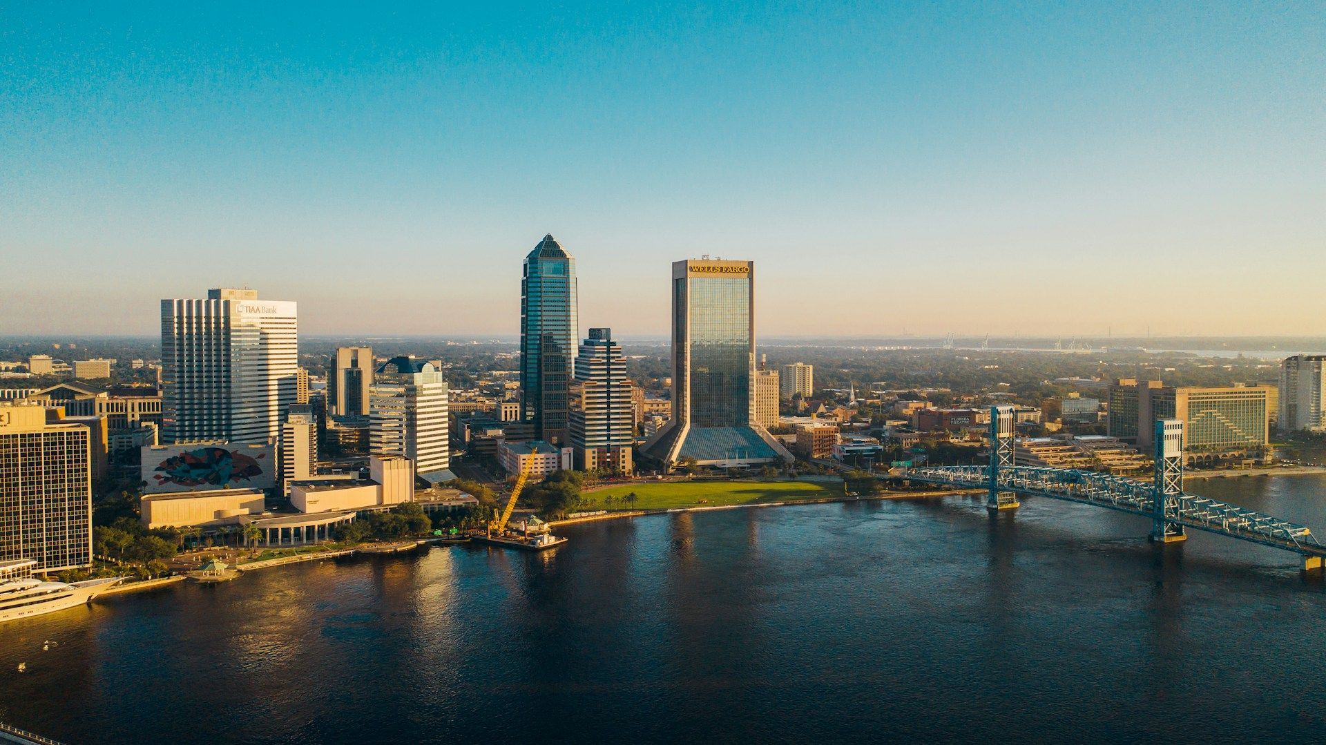 Aerial view of a city skyline beside a river at sunset, with bridges and modern high-rises.