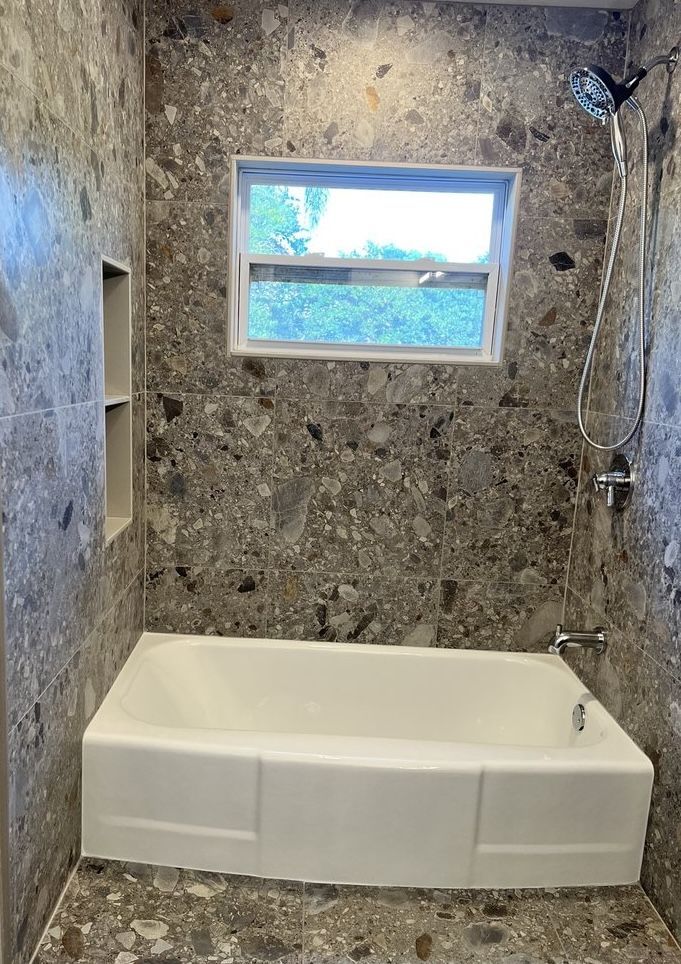 Small stone-tiled bathroom with a white soaking tub, window, and showerhead.