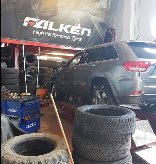 Un SUV grigio con il marchio Falken si trova in un'officina di pneumatici, circondato da pneumatici impilati e attrezzi.