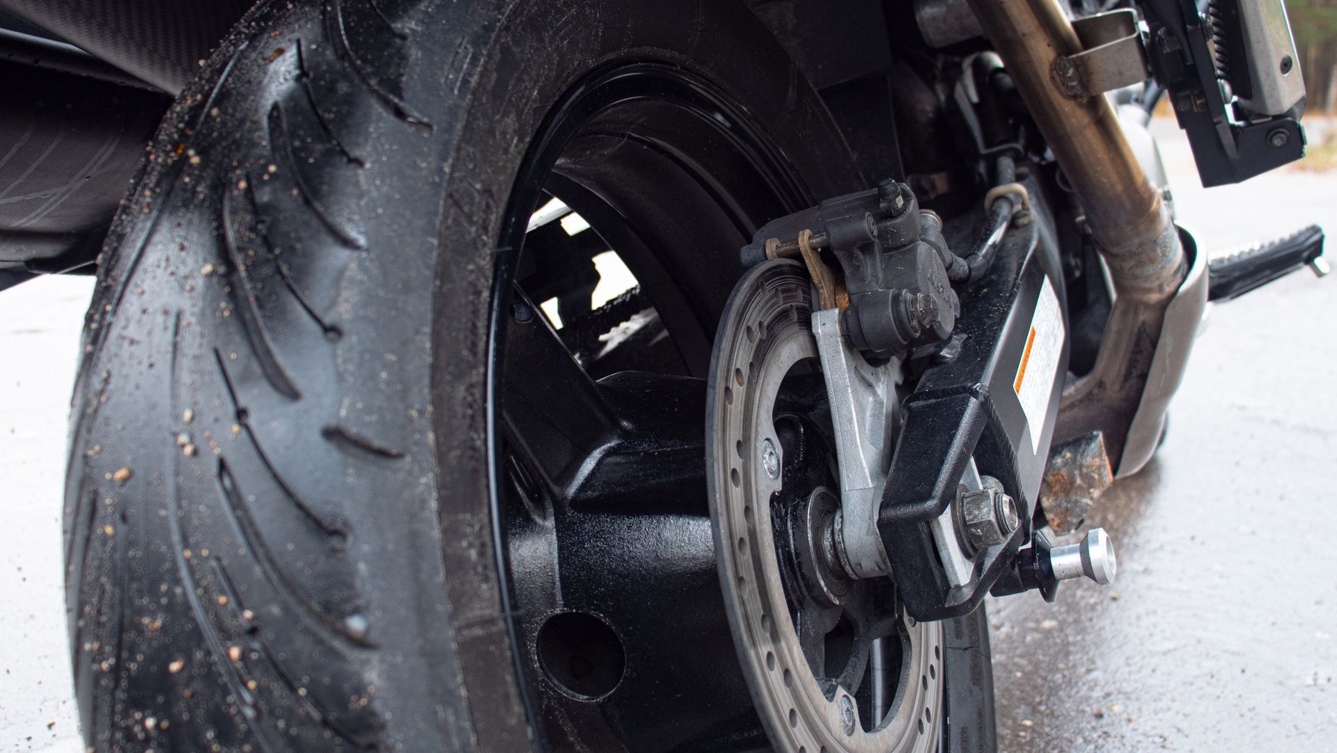 Primo piano di una ruota posteriore e di uno pneumatico di una motocicletta su asfalto bagnato, che mostra il battistrada e i componenti dei freni.