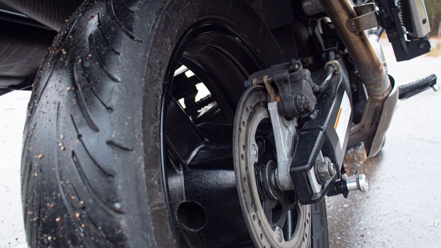 Primo piano di una ruota posteriore e di uno pneumatico di una motocicletta su asfalto bagnato, che mostra il battistrada e i componenti dei freni.