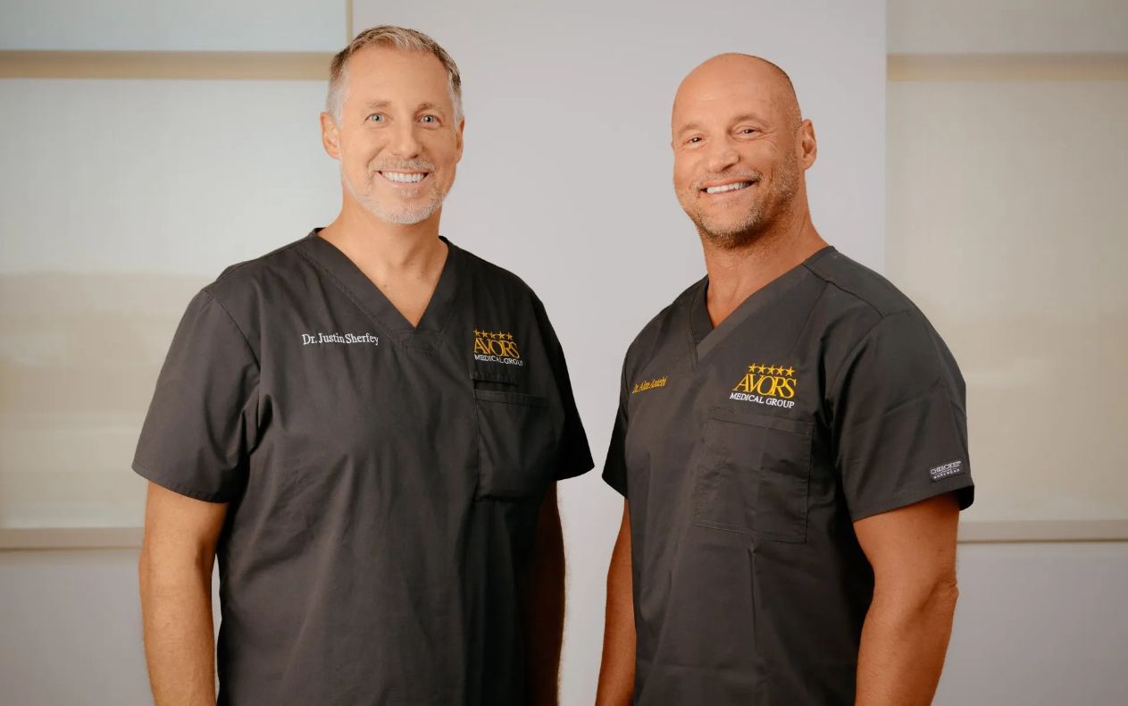 Two men in black polo shirts smiling and standing against a light beige background