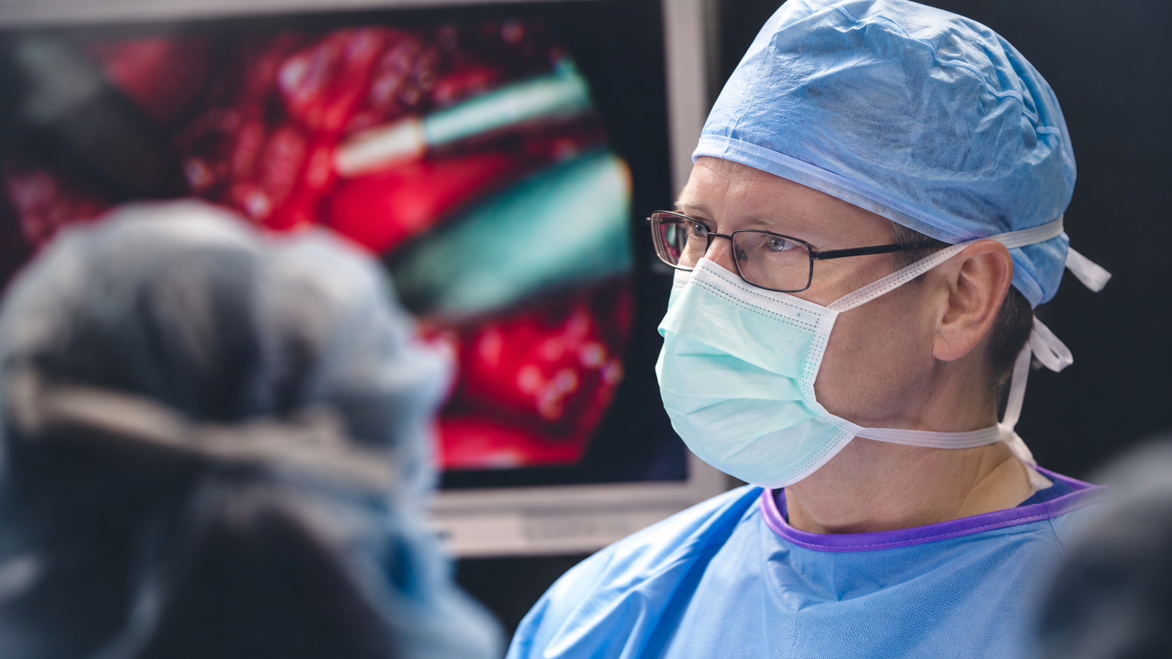 Two surgeons in blue scrubs and masks working in an operating room beside surgical equipment.