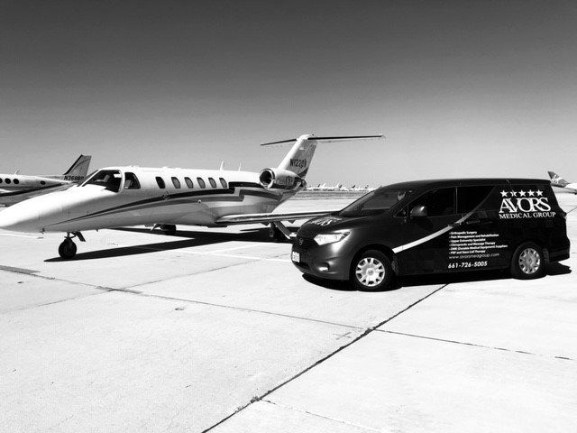 Black and white view: Private jet and branded black van on a tarmac. The van has text reading