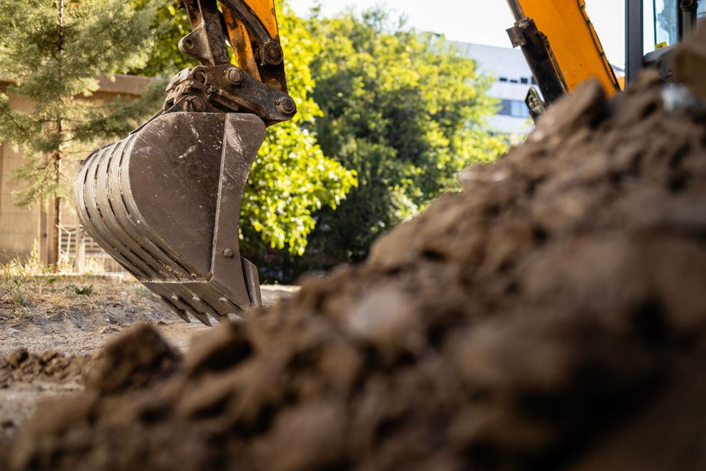 Excavator bucket digging soil at a construction site with trees in the background