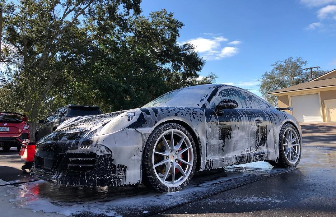 Black sports car covered in soap suds being washed outdoors.