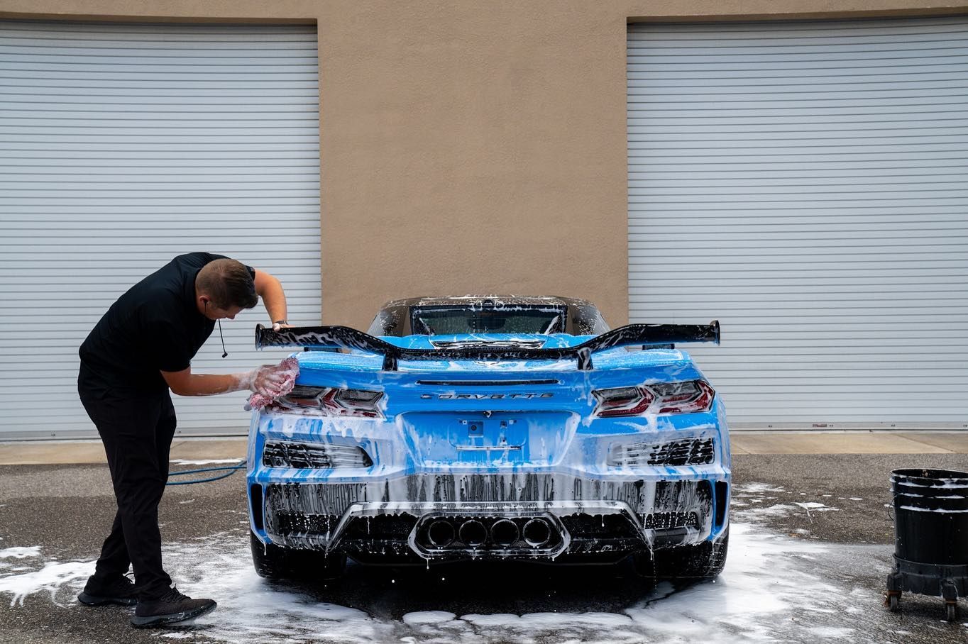Man washing a bright blue sports car outside a garage; soapy water covers the car.