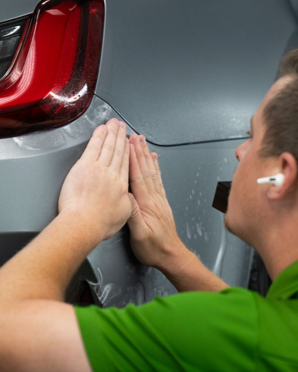 Person sanding a gray car panel near the taillight in an auto body shop