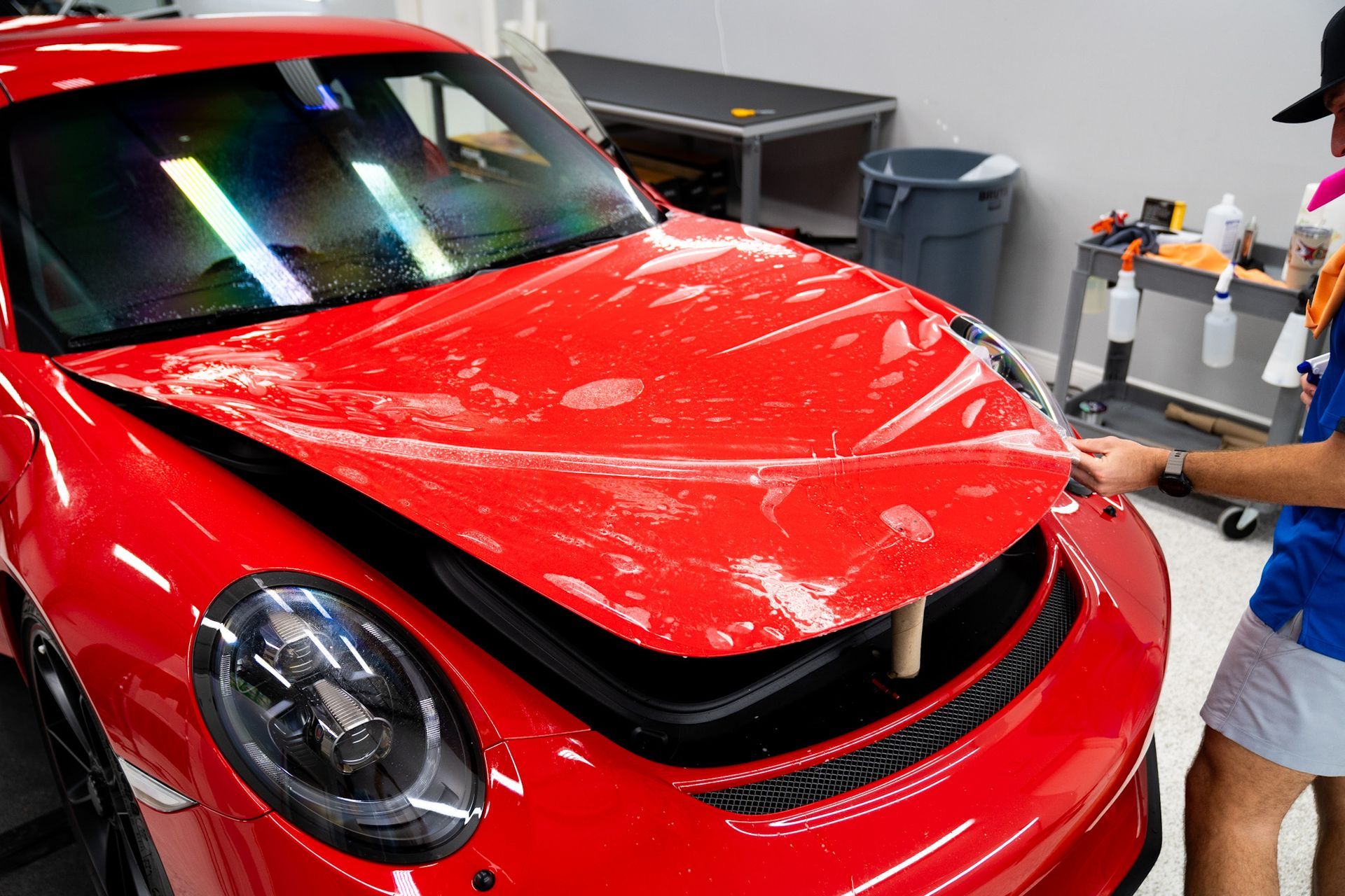 Red sports car hood being covered with clear film by a person in a shop.