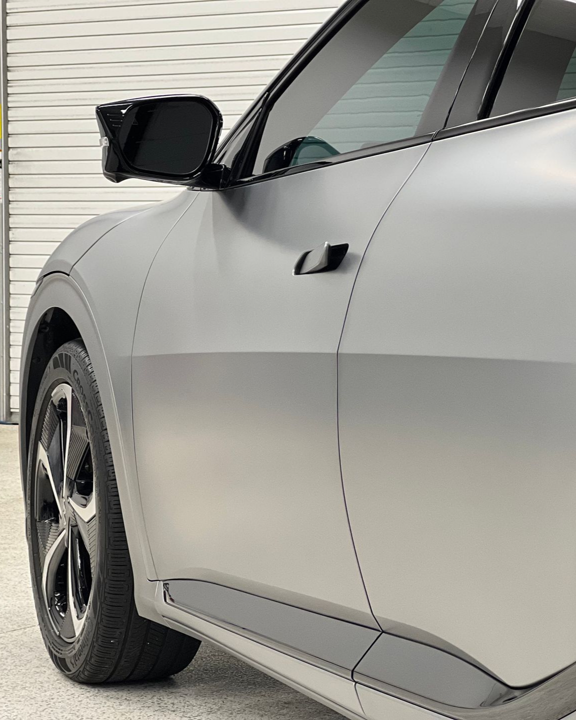 Silver car side view with black wheel and side mirror, parked indoors near a white garage door