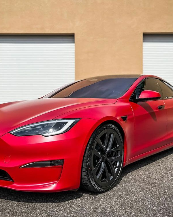 Red Tesla sedan parked outside a beige building, shown from the front side with black wheels.