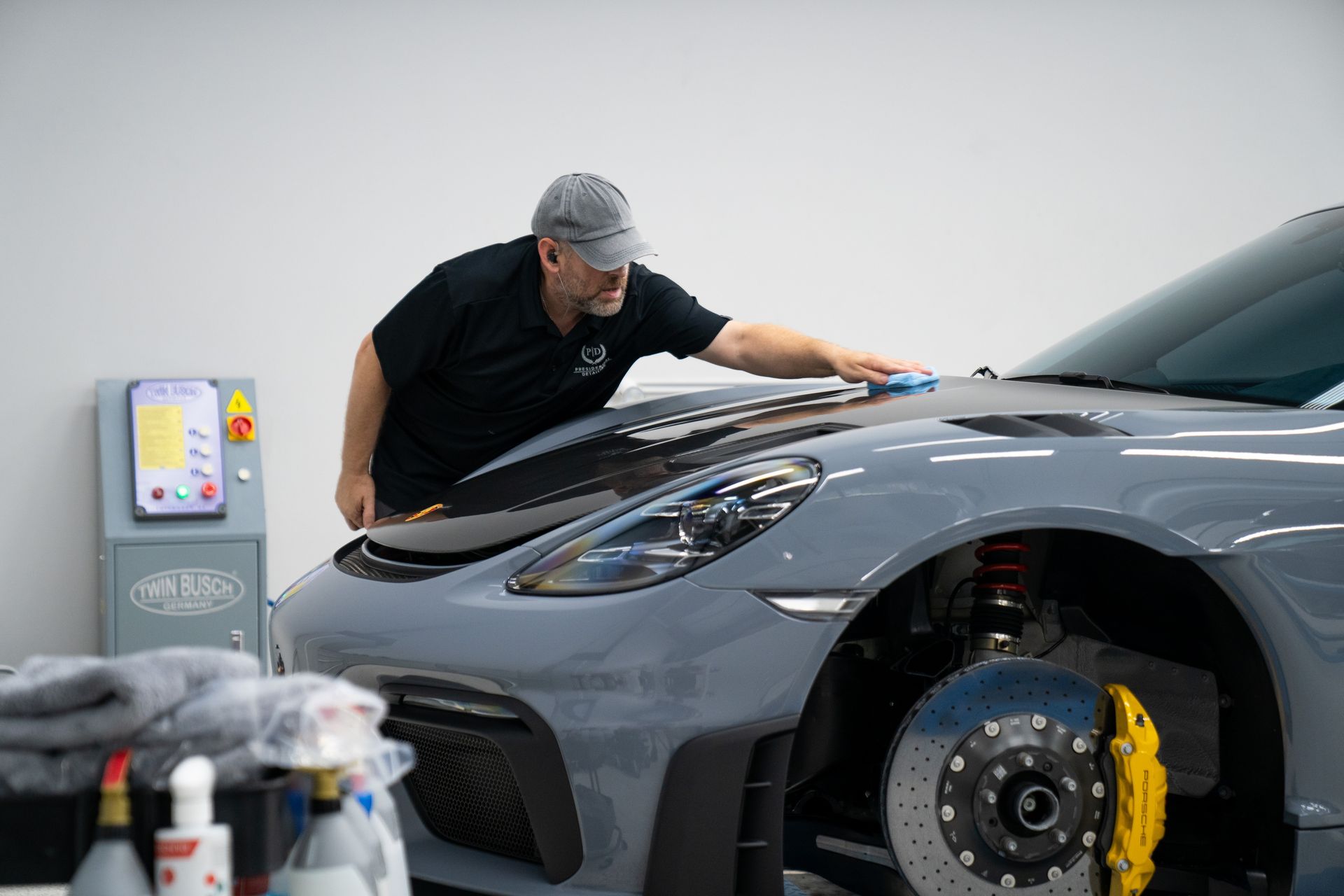 Man polishing a grey sports car's hood in a workshop. Yellow brake calipers, equipment on a nearby table.