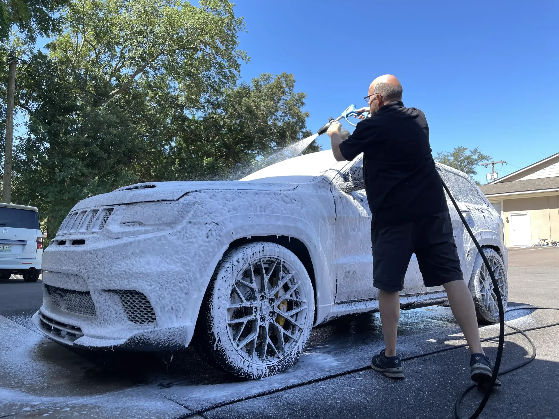 Man washing a white SUV covered in foam outdoors on a sunny day.