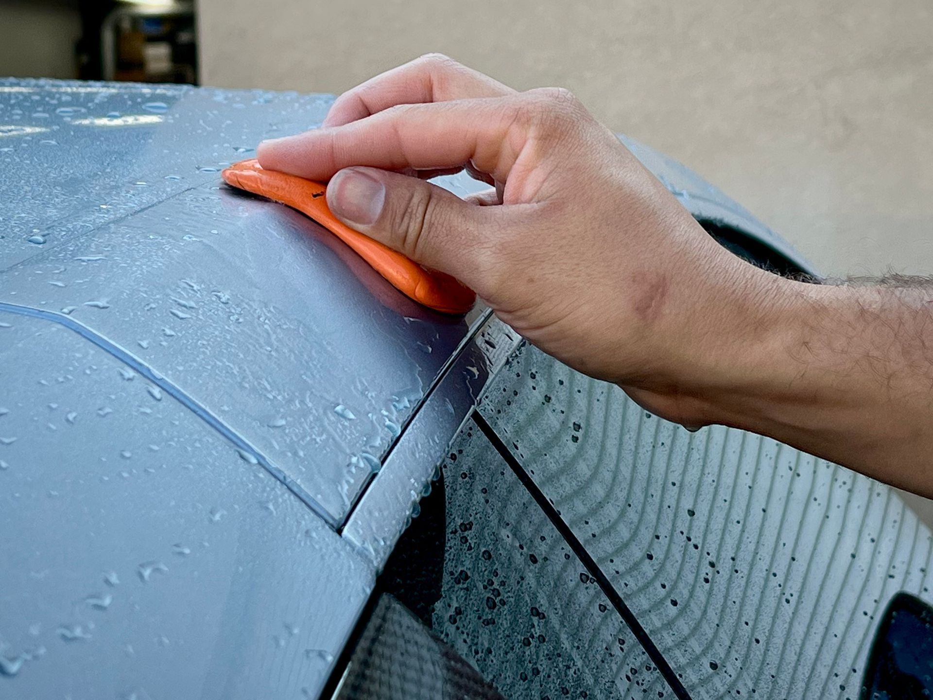 Hand using an orange clay bar on a wet, gray car surface for detailing.