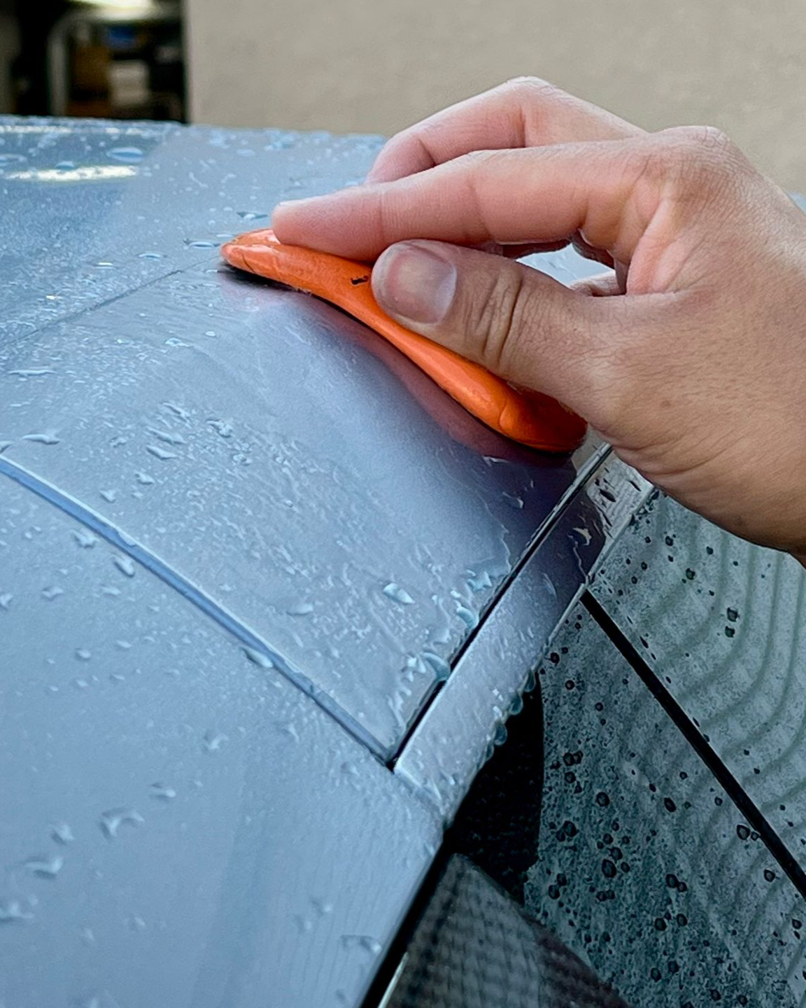 Hand using an orange squeegee on a wet blue car hood during cleaning
