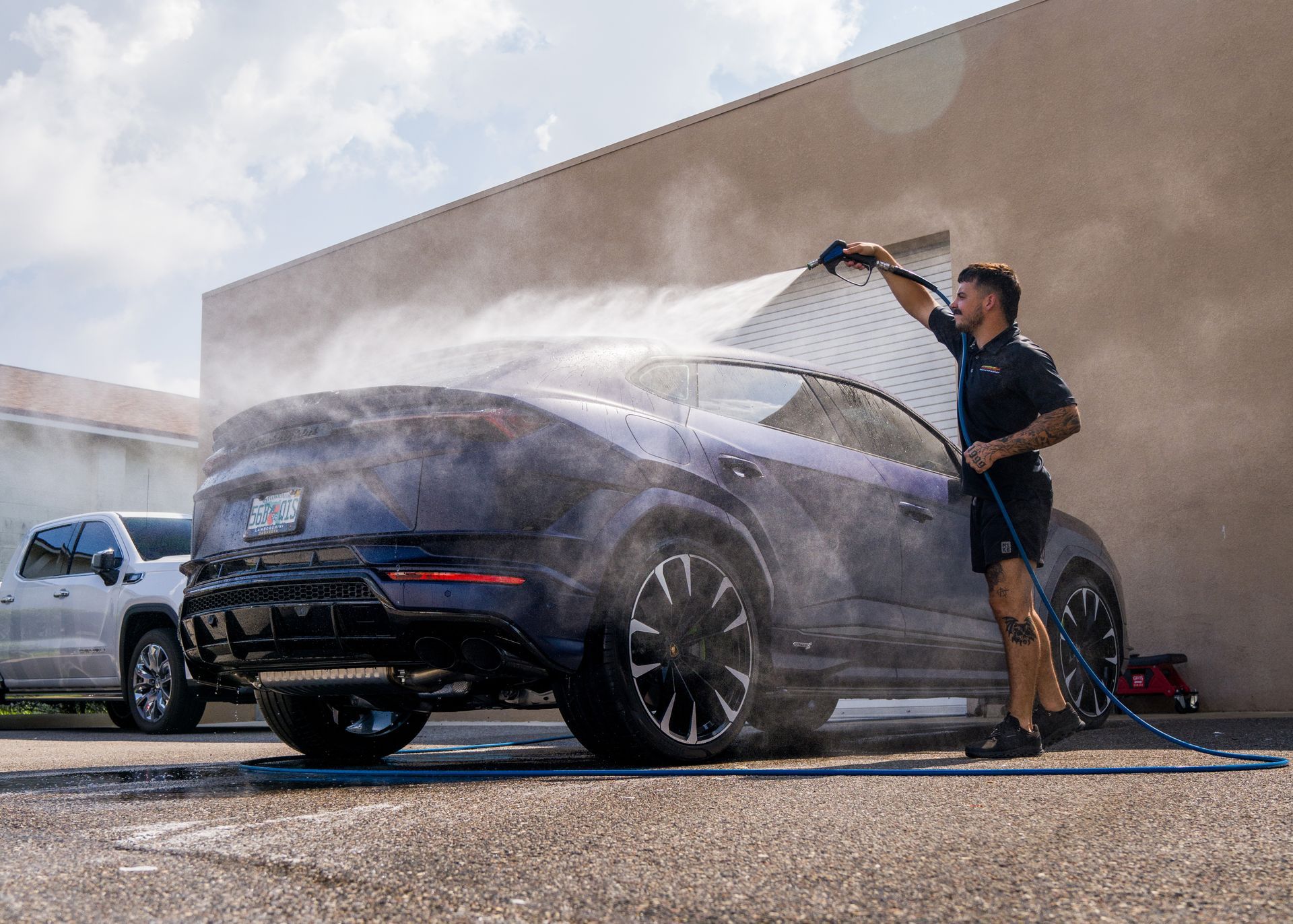 Person pressure-washing a black sports car outdoors, with water spraying across the vehicle