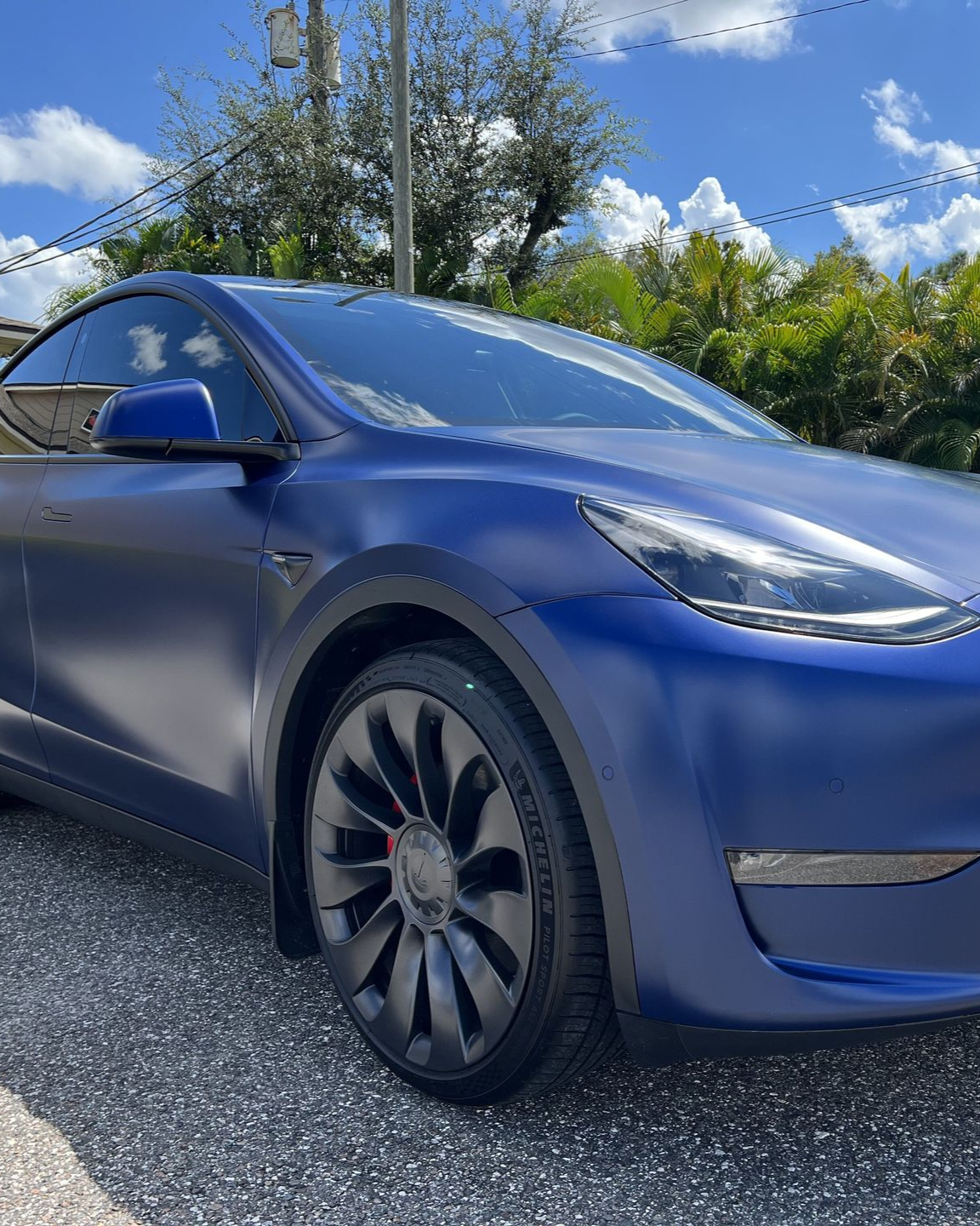 Blue Tesla sedan parked on gravel under a sunny sky with trees in the background