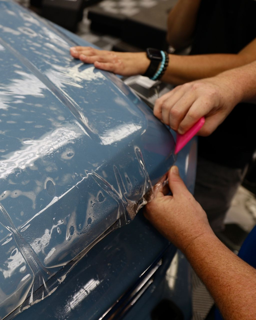 Man applying clear protective film to the front fender of a white car. He is kneeling, working on the wheel well.