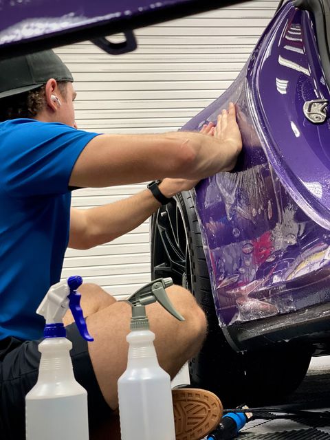Person polishing a purple car panel in a garage, with spray bottles in the foreground