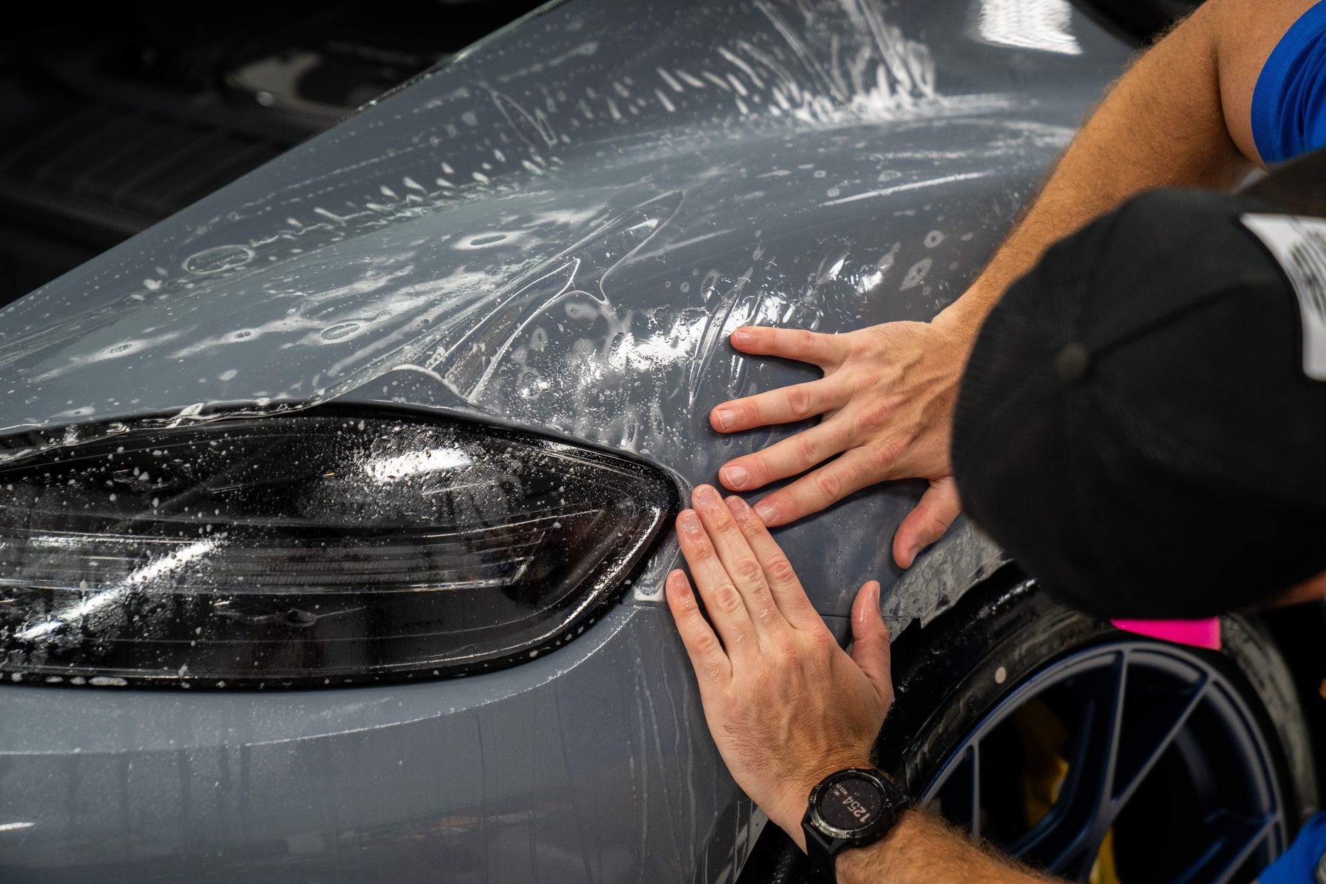 Person applying clear film to a car, using hands to smooth it over the gray hood and headlight.