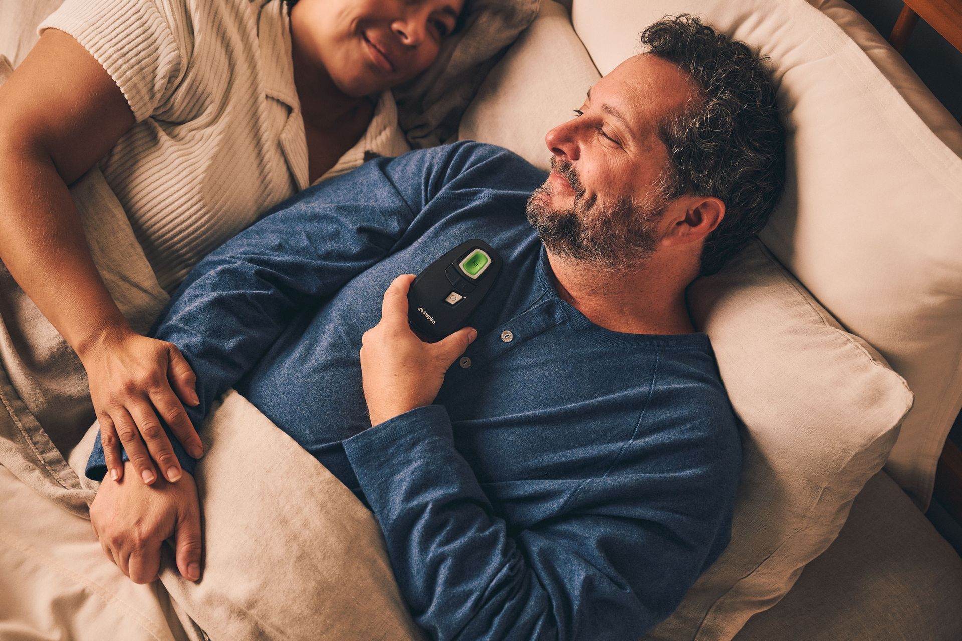Man in bed holding device, woman beside him smiling. Soft lighting, indoor setting.